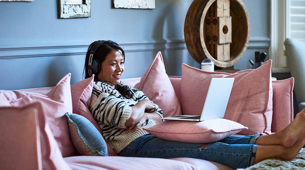 Woman sat on a pink sofa with headphones on and a laptop resting on her lap watching something